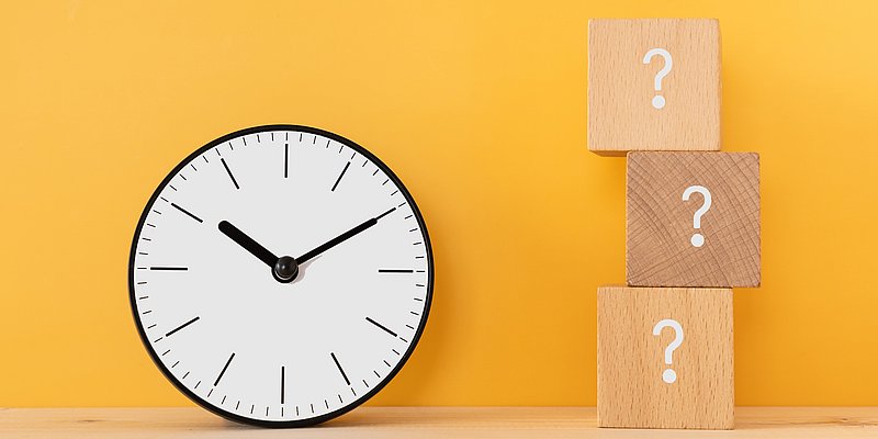 Drei Würfel mit aufgemalten Fragezeichen stehen neben einer Wanduhr