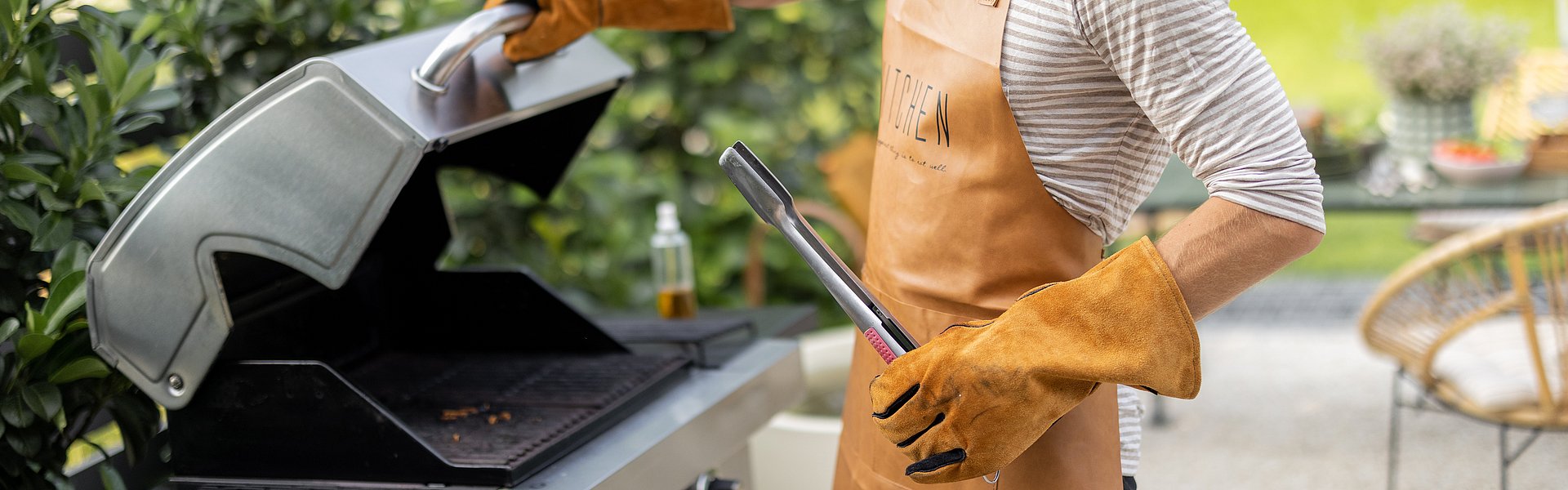 Person steht am Gasgrill mit Grillschürze und Grillzange