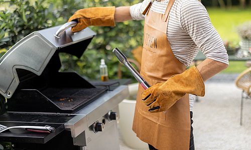 Person steht am Gasgrill mit Grillschürze und Grillzange