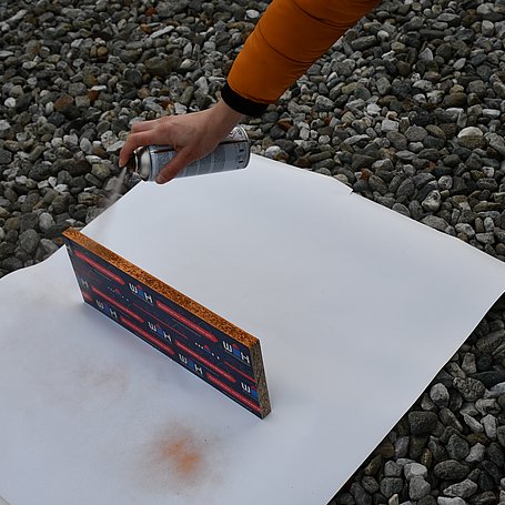 Person sprayt mit einer Sprühdose die Kanten eines Holzbrettes draußen an