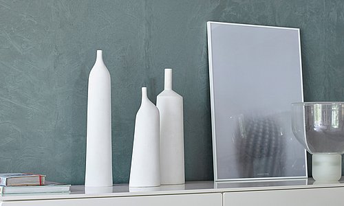 Sideboard an grüner Wand mit weißen Kerzen und roas Blumen als Deko