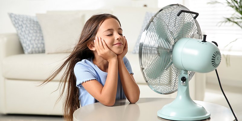 Mädchen sitzt im Wohnzimmer vor einem türkisen Ventilator